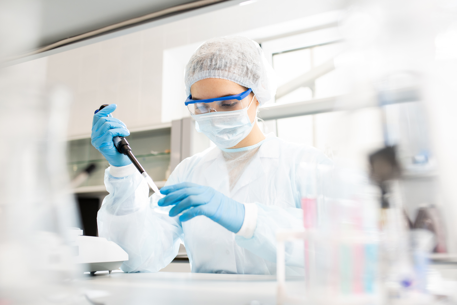 Eine weibliche Laborantin in Schutzkleidung verwendet eine Pipette, um eine Flüssigkeit in ein kleines Gefäß zu übertragen. Sie trägt eine Schutzbrille und eine Maske und arbeitet konzentriert in einem modernen Laborumfeld.