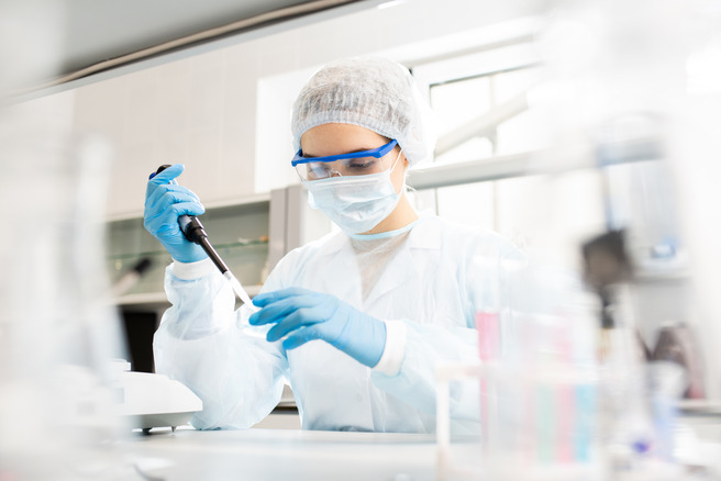 Eine weibliche Laborantin in Schutzkleidung verwendet eine Pipette, um eine Flüssigkeit in ein kleines Gefäß zu übertragen. Sie trägt eine Schutzbrille und eine Maske und arbeitet konzentriert in einem modernen Laborumfeld.