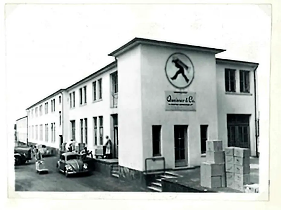 Ein historisches Fabrikgebäude mit dem Logo eines gehenden Mannes. Auf dem Gelände stehen mehrere Personen und Autos, während Kartons gestapelt sind. Die Fassade ist hell, mit mehreren Fenstern und offenen Türen.