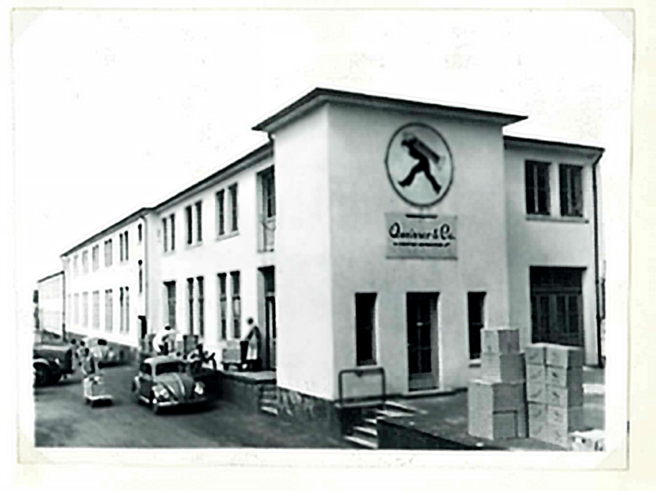 Ein historisches Fabrikgebäude mit dem Logo eines gehenden Mannes. Auf dem Gelände stehen mehrere Personen und Autos, während Kartons gestapelt sind. Die Fassade ist hell, mit mehreren Fenstern und offenen Türen.