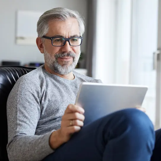 Ein älterer Mann mit Brille sitzt entspannt in einem Sessel und schaut auf ein Tablet. Er trägt ein graues Sweatshirt und hat einen Bart. Die Umgebung wirkt hell und modern.