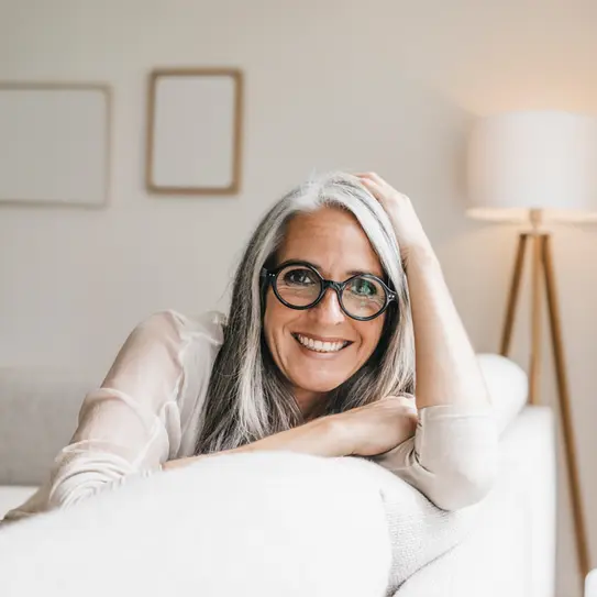 Eine lächelnde Frau mit grauen Haaren und einer runden Brille sitzt auf einem Sofa.