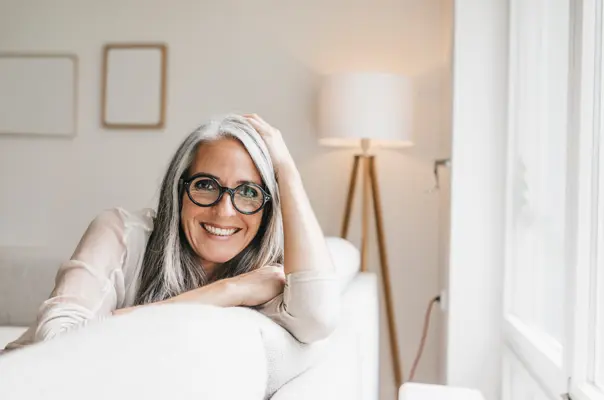 Eine lächelnde Frau mit grauen Haaren und einer runden Brille sitzt auf einem Sofa.