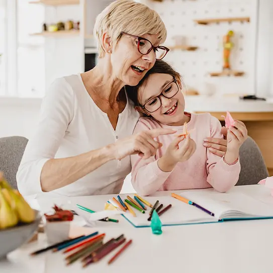 Eine ältere Frau und ein kleines Mädchen sitzen an einem Tisch und gestalten mit Buntstiften und Papier. Die Frau zeigt dem Mädchen etwas, während beide lächeln. Auf dem Tisch sind verschiedene Schreibutensilien und ein Notizbuch zu sehen.