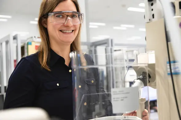 Eine lächelnde Frau in Schutzbrille steht an einem Laborgerät. Im Hintergrund sind Regale mit Laborutensilien zu sehen. Die Szene vermittelt eine positive und konzentrierte Arbeitsatmosphäre im wissenschaftlichen Umfeld.