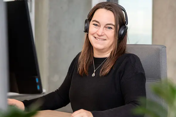 Eine lächelnde Frau trägt Kopfhörer und sitzt an einem Schreibtisch vor einem Computermonitor. Sie hat lange braune Haare und trägt ein schwarzes Oberteil. Die Umgebung wirkt modern und aufgeräumt.