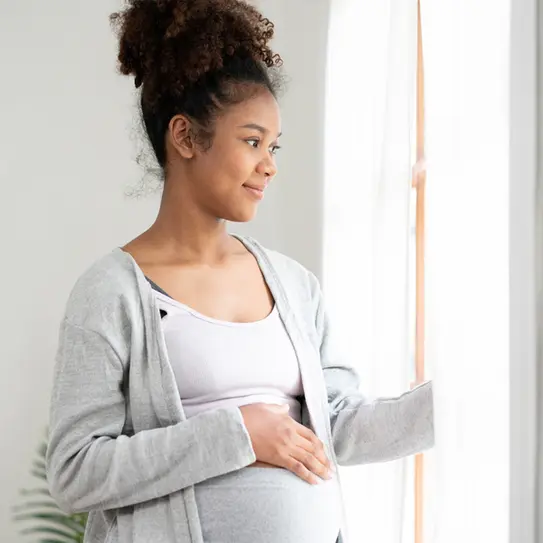 Eine schwangere Frau mit lockigem Haar steht am Fenster und lächelt, während sie ihren Bauch sanft berührt. Sie trägt ein graues Oberteil und eine helle Bluse. Das Licht strömt durch die Fenster und schafft eine warme Atmosphäre im Raum.