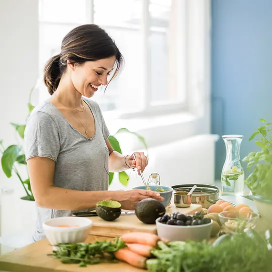 Eine Frau in einem hellen Raum bereitet in einer modernen Küche eine Mahlzeit vor. Sie lächelt, während sie eine Schüssel mit Zutaten umrührt. Auf dem Tisch liegen frisches Gemüse, Avocados und Obst. Pflanzen im Hintergrund verleihen dem Raum eine frische Atmosphäre.
