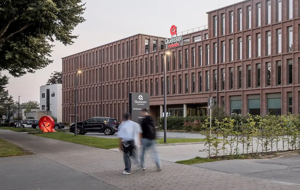 Das Bild zeigt ein modernes Gebäude mit einer roten Ziegelfassade und großen Fenstern, das die Aufschrift „Quasser“ trägt. Im Vordergrund sind zwei Personen zu sehen, die auf einem Gehweg entlang des Gebäudes gehen. Es gibt auch einige Autos und Pflanzen in der Umgebung.
