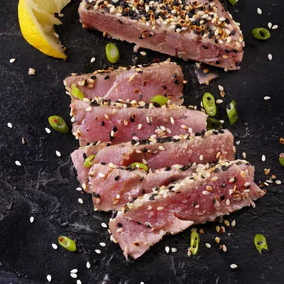 Auf einem schwarzen Plattenteller liegt dünn geschnittenes, leicht angebratenes rotes Fleisch, das mit Sesam bestreut ist. Neben dem Fleisch befinden sich grüne Zwiebelringe und ein Stück Zitrone.