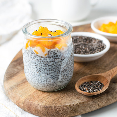 Ein Glas mit Chia-Pudding, garniert mit Würfeln von exotischer Frucht, steht auf einem Holzbrett. Neben dem Glas sind kleine Schalen mit Chiasamen und einer weiteren Obstsortierung. Der Hintergrund ist hell und einfach gehalten.
