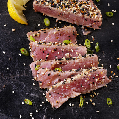 Auf einem schwarzen Plattenteller liegt dünn geschnittenes, leicht angebratenes rotes Fleisch, das mit Sesam bestreut ist. Neben dem Fleisch befinden sich grüne Zwiebelringe und ein Stück Zitrone.