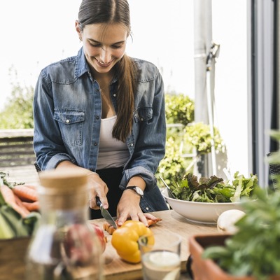 Eine junge Frau schneidet Gemüse auf einem Holztisch. Sie trägt ein Jeanshemd und lächelt, während sie eine gelbe Paprika und andere frische Zutaten wie Salat und Karotten vorbereitet. Im Hintergrund sind Pflanzen sichtbar.