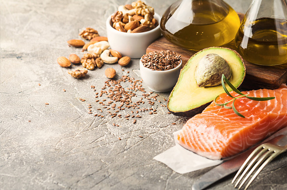 Ein Teller mit frischem Lachs, einer halben Avocado, Nüssen, Leinsamen und zwei Flaschen Olivenöl auf einer grauen Oberfläche.
